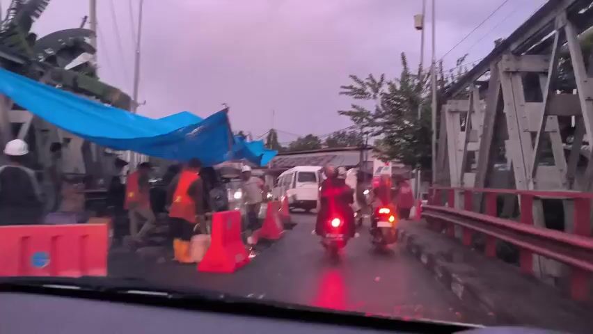 The video shows a driver's perspective of a road scene. The weather appears overcast. Workers in orange vests are congregated under a blue tarp structure on the left side of the road, next to orange traffic barriers. Several motorcycles are driving on the road, one with a red taillight prominently visible. A white van is stopped ahead. A bridge structure is visible on the right side. Inside the car, someone is holding a smartphone and recording the scene through the windshield. The smartphone screen shows a mirrored image of the ongoing recording.