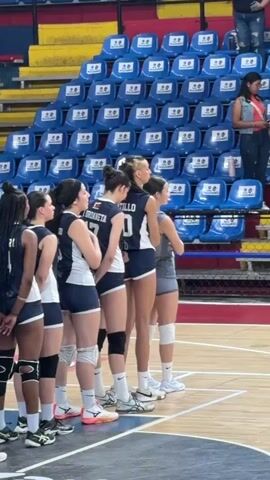 The video shows a volleyball team of young women in navy blue and white uniforms, standing in a line on a wooden court. They are facing away from the camera, toward a set of blue stadium-style seats, most of which are empty. The players have various hairstyles, including braids, ponytails, and buns. Some are wearing knee pads. The court is well-lit, and a referee can be seen walking in the background. There is music playing loudly in the venue.