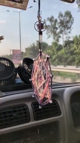 The video shows a view from inside a car driving on a highway. In the foreground, a decorative woven hanging object is visible, along with two black car fans mounted on the dashboard. The windshield is slightly dirty, with streaks. Beyond the windshield, the road is visible with other vehicles, including a white SUV and a truck. Trees and power lines line the side of the road, and the sun is shining brightly in the sky, creating a lens flare effect. The overall environment appears to be daytime, possibly in a rural or semi-urban setting.