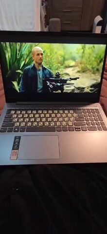 A laptop sits on a dark, soft surface, likely a bed. The screen displays a scene from a film or show, transitioning between male and female actors in outdoor settings. The laptop is silver with a standard keyboard featuring both Latin and Cyrillic characters, covered with yellow stickers for the Cyrillic layout. A small set of manufacturer's stickers is near the trackpad. The background beyond the laptop includes glimpses of a wooden cabinet, a white blanket, and a decorative object.