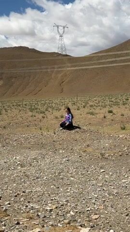 A young woman with long dark hair, wearing a colorful tie-dye long-sleeve shirt, black pants, and white shoes, is seated cross-legged on the ground in a wide shot. She faces forward and slightly to her left, with a calm and contemplative expression. The setting is an arid, sparsely vegetated landscape, possibly a high-altitude or plateau region, with dry earth, rocks, and scattered small plants. In the background, a large, barren brown hill or mountain dominates the horizon. A prominent electricity pylon (power line tower) stands tall on the distant hillside, with power lines stretching across the mid-ground. The sky above is bright with scattered white clouds, indicating daytime. The scene conveys a sense of peaceful solitude in a remote, natural environment, consistent with the Mainpu township, China location context. There is no visible text in the video.