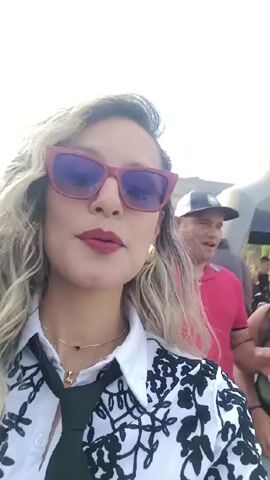 A woman with long, wavy blonde hair and red-framed sunglasses smiles at the camera in a selfie video. She wears a white blouse with black floral embroidery and a black tie. She has red lipstick and gold earrings. Behind her, there is a crowd of people, including men in casual clothing.  A large green balloon with a logo is visible, as well as promotional tents, motorcycles, and inflated tires. The setting appears to be an outdoor event with various vendors and attendees. The weather looks sunny and the atmosphere lively.