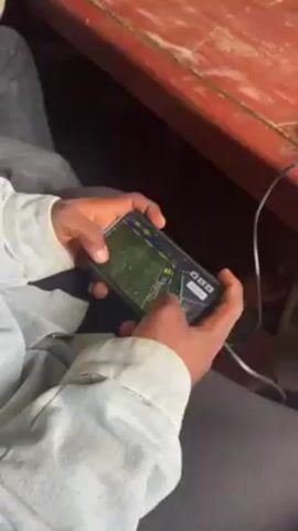 The video shows a person playing a soccer video game on their smartphone. The person is seated at a desk in a classroom filled with other students. The phone is held horizontally, and the person uses both thumbs to control the game. The classroom appears to be somewhat crowded, with students seated at rows of desks. The environment is lit by fluorescent lights.