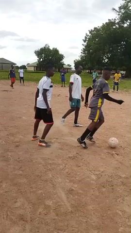 The video shows a group of young males playing soccer on a dirt field. In the foreground, three males are running, one of whom is kicking a soccer ball. In the background, several other males are observing or participating in the game. There is a large tree in the middle ground and a building behind it. The sky is overcast.