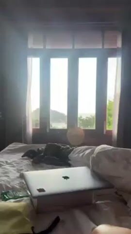 The video shows a bedroom in what appears to be a tropical setting. A large bed with white sheets is in the foreground, and on it, there is a closed laptop, a small pillow, and some crumpled clothing.  In the background, a large window with four panes offers a view of the ocean, lush green trees, rooftops, and a small island in the distance. A straw hat hangs on the window frame. The room has wooden floors and dark curtains. The light is bright, entering through the window.