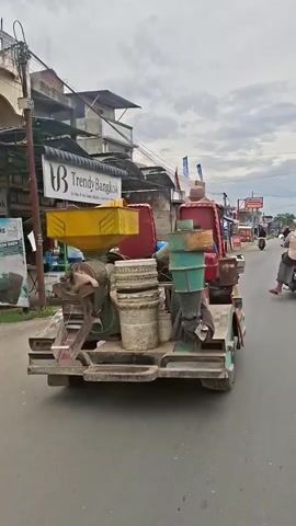The video shows a three-wheeled vehicle loaded with various items, including buckets, containers, and what appears to be machinery. The vehicle is on a paved road, with buildings and shops visible in the background. The sky is overcast. A person can be seen walking in the distance on the right side of the frame. The camera is positioned behind the vehicle, capturing a rear view. The vehicle appears to be moving.