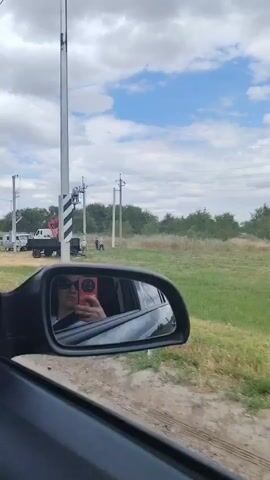 The video pans across an outdoor scene. In the foreground, the reflection of a person holding a red smartphone and wearing sunglasses is visible in a car's side mirror. The environment is a grassy field with some dry patches. In the background, several utility poles are present, with one having a crane extending towards it. A white truck with a crane is parked near the utility poles, and a small grey van is also visible. Another utility pole is shown in closer detail towards the right side of the frame. Trees line the horizon. The sky is cloudy with patches of blue.