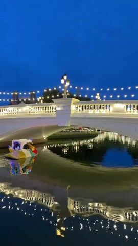 A video shows a canal at dusk. A white bridge with lampposts and string lights spans the canal. On the left side of the canal, there is a small, colorful boat decorated with lights. The boat has two figures in it. The water reflects the lights from the bridge and the boat. In the background, there are buildings and trees. On the right side of the canal, there is a brick lighthouse.