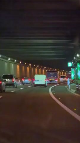This video captures traffic flow within a brightly lit tunnel, likely a highway or major road. The perspective is from inside a vehicle, looking forward at the road ahead and other vehicles.

**Subjects:** The primary subjects are vehicles, including cars, a white van, and a larger bus. There are no visible people.

**Activity/Event:** The activity is the movement of traffic through the tunnel. The vehicles appear to be moving at a moderate pace, with some congestion visible.

**Setting:** The setting is a tunnel, characterized by its artificial lighting along the walls and ceiling. The road surface is dark asphalt, marked with white lane lines and reflective road studs. The tunnel ceiling has a ribbed, industrial appearance.

**Notable Details:**
*   **Time of Day:** The interior lighting and the presence of headlights on vehicles suggest it is either nighttime or the tunnel is continuously illuminated.
*   **Weather:** The weather is not discernible from the interior of the tunnel.
*   **Location Cues:** A green exit sign with white text and an arrow is visible on the right side of the tunnel, indicating an upcoming exit. Another sign with blue and white lettering is also present. The location context provided is Sant Joan D'alacant, Spain.
*   **Emotions:** No specific emotions are conveyed due to the absence of human subjects and the focus on vehicular traffic.

**Visible Text:**
*   "EXIT" is visible on the green sign on the right, along with an arrow.
*   The license plate of the white van is partially visible, appearing to be European in format (e.g., a series of letters and numbers).
*   Text is present on the back of the blue bus, but it is too small and blurred to be legible.
*   Some text is visible on signs on the right side of the tunnel, but it is not clearly discernible.