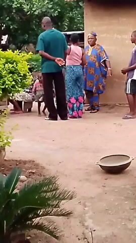 A group of people are gathered in an outdoor, possibly rural, setting. The ground is dirt and there is vegetation visible. A building with a corrugated metal roof is in the background. In the foreground, a young child walks from left to right. To the left of the child, an adult man in a green shirt and black pants stands with his back to the camera, facing a woman in a pink top and patterned pants. To the right of the woman, an adult man in a blue and gold patterned robe and a blue cap is facing the woman and man. Further to the right, a young person in a purple and white shirt and black shorts stands looking towards the other individuals. A large, shallow metal bowl is on the ground in the right foreground.