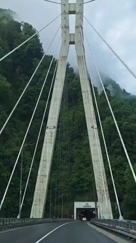 The video shows a low-angle shot of a cable-stayed bridge. The primary visual focus is on the central support tower and the cables extending from it. The tower is made of concrete and has a distinctive "H" shape at the top. Several white cables are strung from the tower diagonally, supporting the bridge deck below. Additional black cables run vertically down from the bridge. In the background, a lush green mountain landscape is partially obscured by low-hanging clouds. The sky transitions from a bright blue to darker, stormy-looking clouds. Music plays throughout the video.