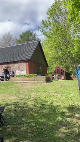 The video shows an outdoor gathering of people on a sunny day with partly cloudy skies. The setting appears to be a park or a large yard with a building in the background made of stone and brick. Some people are sitting at picnic tables near the building, while others are standing in groups on a grassy area and a dirt path. People can be seen eating, adjusting their gear, and interacting with each other. Some are wearing brightly colored vests, and at least one person has a backpack. The environment includes grass, trees, and a small red shed-like structure.