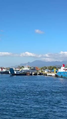 The video shows a wide shot of a harbor on a clear day with a bright blue sky. Several ferries are docked at a pier. In the foreground, the blue water of the harbor is visible with gentle waves. Two large ferries are prominently featured. One is a blue and white ferry with red accents on top. The other is a white ferry with blue trim and the text "The Spirit of Journey" visible on its side. The ferries are connected to the pier by ramps. The pier itself is lined with buildings with blue roofs and yellow structures. In the background, lush green mountains rise against the sky. Two tall white towers, possibly communication towers, are visible near the shore. The overall scene depicts a busy ferry terminal with boats ready for departure or arrival. There are no people visible in the frame.