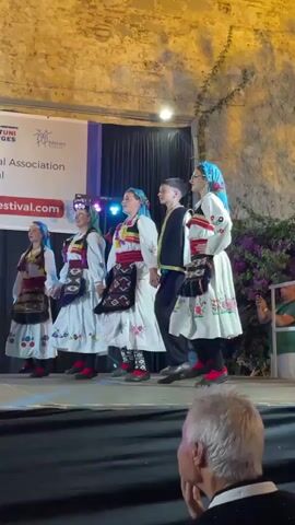 This is a video of a performance at the Calella Folklore Festival in Calella, Spain. Five individuals, four females and one male, are on stage dancing. The four females are dressed in traditional folk costumes, consisting of white dresses with embroidered details and colorful aprons adorned with geometric patterns. They also wear headscarves. The male performer is dressed in a dark vest over a white shirt, dark pants, and dark shoes.

The stage is set up outdoors, with a backdrop featuring banners with text. One banner reads "Ayuntament de Calella" and "FORTUNI VIATGES" and "Folklore Festival Association". Another banner prominently displays "Calella Folk Festival" in large red lettering. The ground behind the performers is dark, and the performers are on a raised stage with a bright lighting setup. The background wall appears to be made of stone. In the foreground, the back of a spectator's head, wearing a white baseball cap, is visible. The lighting suggests it is likely evening or nighttime. The performers appear engaged and are executing synchronized dance moves. The atmosphere seems festive and cultural.