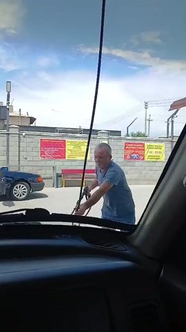 A man is seen at a car wash, holding a pressure washer hose, appearing to be washing a blue sedan. The car's doors are open, and there seem to be two people inside the vehicle. The man is wearing a grey t-shirt and jeans, and he is focused on the task of washing the car. The environment appears to be an outdoor car wash facility, with a concrete ground and a brick wall in the background. The sky is clear and blue, suggesting a sunny day. The video is shot from inside another vehicle, likely the one being washed, looking out through the windshield.