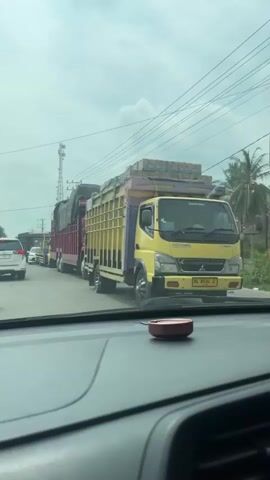 The video shows a line of trucks on a road. The camera is inside a car, looking out at the traffic. The trucks are large and appear to be carrying goods. The sky is cloudy. There is a white SUV in front of the car, and other vehicles can be seen further up the road. The trucks are of various colors, including red, yellow, and purple. One of the trucks has a tarp covering its load. There is a cell tower in the background. The overall scene suggests a traffic jam.
