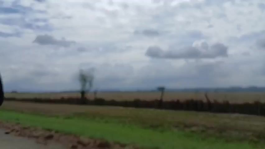 The video shows a view from a moving vehicle, likely a car, driving alongside a field. The sky is visible, filled with white clouds. The field, mostly brown, appears to be harvested or fallow. A sparse line of vegetation divides the road and the field. In the far distance, hints of what could be mountains are visible. The overall quality of the video is somewhat shaky and slightly out of focus. The sound suggests wind noise is present.