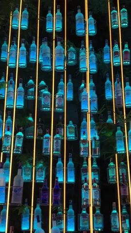 This media file depicts a wall of bottles displayed in a dimly lit setting, possibly a bar or nightclub in Baan Lamai, Thailand. The bottles are of various shapes and sizes, and many are illuminated with blue and yellow lights, creating a vibrant and atmospheric display. Some bottles appear to contain liquids, while others seem to be empty. The bottles are arranged in vertical rows, with thin yellow bars interspersed between them, providing a structural and decorative element.

The scene is dark, suggesting it is nighttime. There is no visible weather information. The presence of multiple Coca-Cola branded refrigerators at the bottom of the frame indicates a commercial establishment that serves beverages. There are no clearly identifiable people in the foreground, though shadows and glimmers suggest the possibility of patrons or staff in the periphery. The overall impression is one of a lively and visually engaging environment, likely designed to attract customers and enhance the ambiance. The blue and yellow lighting creates a modern and somewhat futuristic feel.