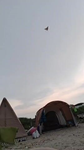 The video shows two tents set up on a sandy surface, likely a beach, under a cloudy sky. A kite is visible flying in the upper center of the frame. The tents are of different sizes and colors; one is a light brown triangular tent, and the other is a larger, darker brown dome tent with its entrance open. Several items are placed between the tents, including bags and possibly camping equipment. In the background, there appears to be a line of vegetation or grass, and a hint of water in the distance. The lighting suggests it might be early morning or late evening. The camera is positioned low, looking upwards towards the sky.
