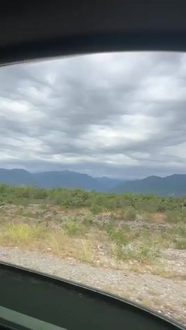 The video shows a view from inside a vehicle, looking out a window at a cloudy sky and a mountainous landscape. The foreground is a gravelly roadside with some sparse vegetation. In the mid-ground, there are green trees and bushes. The background consists of layers of mountains fading into the distance, with a heavy, overcast sky above. The camera appears to be moving, as indicated by the slight blur and shifting perspective of the scene. There are no people or distinct objects visible beyond the natural landscape and the vehicle's window frame. The overall impression is one of a scenic drive through a rugged, natural environment.