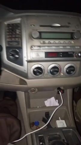 The video captures the interior of a car driving at night in Iyara, Nigeria. The initial view shows the center console with an older model stereo and air conditioning unit, featuring multiple buttons and knobs. A white charging cable and a white USB wall charger are visible in a storage compartment below the dashboard, alongside some small pieces of paper.

The camera then pans to the left, revealing the steering wheel, which has a black and reddish-brown/orange patterned cover. A prominent gold Toyota emblem is centered on the steering wheel. Behind it, the instrument cluster is illuminated, displaying a tachometer on the left, a speedometer in the center, and other gauges on the right. Several yellow and red warning lights are active, including what appears to be the check engine, low fuel, battery, and seatbelt indicators.

Finally, the shot moves to the view through the windshield, showing a very dark, unpaved road ahead, illuminated only by the car's headlights. The overall darkness confirms it is nighttime. Throughout the video, a conversation is audible, with one voice distinctly saying, "We making money." No people are directly visible in the video frame.