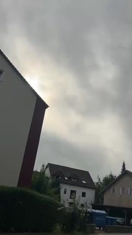 The video begins with a shot of a cloudy sky over houses and trees. The view then shifts to a street lined with houses and parked cars. A blue car is visible in the foreground. The video continues to pan across the street, showing more houses and a church tower in the distance. Finally, the camera focuses on a street sign that reads "Karl-Marx-Weg" and a pedestrian crossing sign.