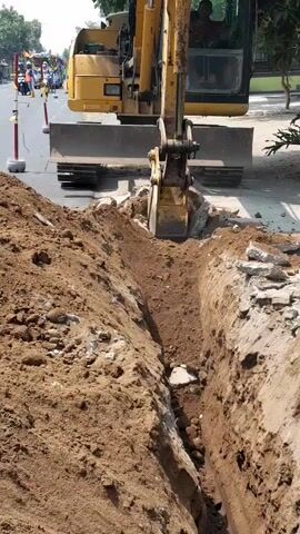 An excavator is digging a trench in the ground. The excavator is yellow and has a large bucket at the end of its arm. The trench is narrow and deep, and there is loose dirt and rocks around it. In the background, there is a road with traffic and some people. The scene appears to be a construction site.