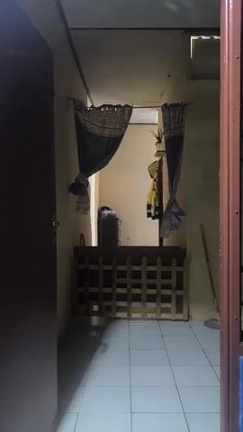 This is a first-person perspective video of an indoor hallway. The camera is positioned in the center of the hallway, facing forward. The hallway is narrow and appears to be made of concrete walls and a tiled floor. There are two doorways visible, one on the left and one on the right. The doorway on the left is open and shows a dark interior. The doorway on the right is partially visible and appears to be a darker color.

In the center of the hallway, there is a wooden fence-like structure. Behind the fence, there are two curtains hanging from the ceiling. The curtains are dark in color and have a patterned trim. Behind the curtains, there is a glimpse of a room with a dresser and some clothes hanging on the wall. There is also a broom leaning against the right wall. The lighting in the hallway is dim, with some natural light coming from the room at the end of the hallway. The video is static with no movement.