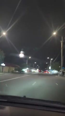 This media file captures a nighttime drive through Parakar, Armenia. The scene is a street illuminated by streetlights and the headlights of passing cars. The camera is positioned inside a vehicle, looking forward through the windshield. The road is asphalt with clear white lane markings.

On either side of the street are buildings, some appearing to be shops or businesses with lights on. Trees are visible along the roadside, their shapes softened by the darkness and artificial light. Several cars are present on the road, including a few in the foreground and others further down the street, some with their brake lights illuminated, indicating traffic or waiting at an intersection.

The time of day is clearly night, with a dark sky overhead. There are no strong indicators of weather conditions, but the lighting suggests it's likely clear. The overall atmosphere is calm and ordinary, depicting a typical late-night drive. There is no visible text in the media file.