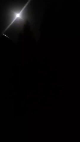 The video shows a dark scene with a bright light, possibly the moon, in the upper left corner. A silhouetted figure of a person is visible standing in front of the light. The rest of the scene is dark and indistinct, with some vague shapes that could be trees or other objects in the background. The person appears to be stationary.
