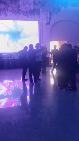 The video shows a group of people dancing in a dimly lit room with purple and pink lighting effects. There is a large screen in the background displaying a rotating circular pattern of red and yellow lights. Several individuals are silhouetted against the screen, their movements illuminated by the lights. The floor appears to be reflective, showing the colorful light patterns.