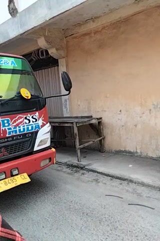A red Mitsubishi truck is parked in what appears to be a repair bay or garage. The truck is tilted, with its front end slightly elevated. A man's face is visible through the driver's side window, looking out. The truck has "BAPUTRI Z&A" and "BOSS MUDA" written on the windshield and the front of the cab, respectively. A yellow license plate with the inscription "BK 8495 CL" is attached to the front bumper. There is a large, black mirror attached to the driver's side door. The floor is concrete, and there are some metal beams and a wooden ladder visible in the background. Underneath the truck, towards the rear, a folded umbrella or tarp is visible.