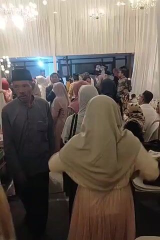 The video shows a group of people at an indoor event, possibly a wedding reception. White curtains hang in the background, with chandeliers visible at the top. In the foreground, several women wearing hijabs are present. One woman in a light brown hijab and a beige dress is facing away from the camera, with her arms outstretched. To her left, another woman in a pink hijab and a patterned dress is visible, also facing away. A man in a dark shirt and a peci hat is in the left foreground, facing mostly towards the camera but with his head turned. Other attendees, some in formal attire, are visible in the background. There are tables with items on them, including what appears to be a gift box with a floral pattern. The overall atmosphere seems festive.