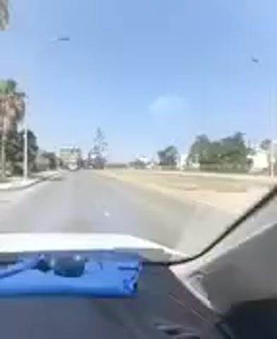 The video shows a view from inside a vehicle, looking out through the windshield. The vehicle appears to be driving on a paved road. The road is bordered by a sandy or dirt shoulder on the right. On the left side of the road, there are trees and some buildings. In the distance, there are more trees and buildings, suggesting an urban or suburban environment. The sky is clear and blue, indicating a sunny day. There are some faint dark objects in the sky, possibly birds or debris. The dashboard of the vehicle is visible in the foreground, with a blue object and some other items on it. The camera is angled slightly downwards.
