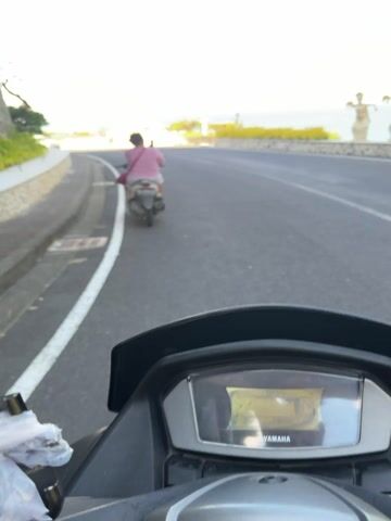 The video captures a first-person view from a moving scooter, following another person on a scooter along a paved road. The road runs parallel to the ocean, which is visible on the right side of the frame under a blue sky with scattered white clouds. To the left, a stone wall topped with lush green plants borders the road, with a large tree growing vertically alongside it.

The person in front is riding a light-colored scooter and wearing a pink top and light-colored shorts. A red bag hangs from their shoulder. The scooter's license plate reads "DK 7860 IC."

As the scooters proceed, a prominent landmark comes into view: two large white swan sculptures, with their necks forming a heart shape. Behind them, a sign on a stone base reads "Blue Ocean Beach UNGASAN." A silver car is also visible near the sign. The scene depicts a scenic coastal drive, likely in the specified location of South Kuta, Indonesia, during the daytime.