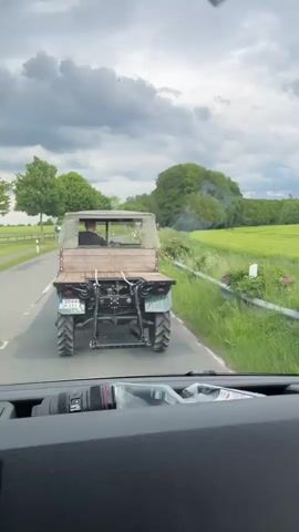 The video shows a view from inside a moving vehicle on a rural road. In the foreground, the dashboard and rearview mirror of the vehicle are visible. Through the windshield, a small vintage tractor is seen driving ahead on the road. The tractor has a flat wooden bed and a driver is visible in the cab. The vehicle filming is overtaking the tractor. Outside the vehicle windows are green fields and trees under a cloudy sky.