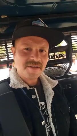 A man wearing a black cap, black jacket, and a mustache sits in the back of an auto rickshaw, holding a camera and facing the viewer. The rickshaw is moving down a street with buildings and shops on either side. The driver is seen in front, wearing a light-colored shirt and a pink scarf around his neck. There are other passengers in the rickshaw. The sound of the horn is audible.