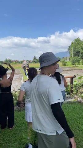 A group of people are standing and walking in an outdoor environment. The foreground shows a grassy area with a person in a bucket hat looking back towards the camera. Behind them, several other individuals are visible, some with their backs to the camera, others looking forward. One person is wearing a black tank top and black pants, another a light green t-shirt and white skirt, and another a white t-shirt and blue jeans. They appear to be on a path or walkway. In the mid-ground and background, there is a field, possibly a rice paddy, with water. There are some structures and vegetation visible, and in the far distance, mountains are present under a blue sky with scattered clouds. The lighting suggests it is daytime.