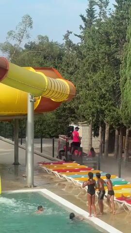 At 00:00, a water slide at a water park is visible, with a yellow and orange tube section at the top and a red and yellow tube section curving downwards. Several people, wearing bright pink shirts, are standing on a platform or cart beneath the slide, seemingly supervising. A few children are in a swimming pool in the foreground. At 00:01, the scene remains the same. At 00:02, one of the individuals in the pink shirts appears to be moving, possibly preparing for a slide. A child in the pool is visible. At 00:03, the water slide continues to be prominent. More people are seen standing near the slide entrance. Children are still in the pool. At 00:04, the perspective widens slightly, showing more of the water park environment. The slide continues to be the main focus. People are still present near the slide, and children are in the pool. The surrounding area is lush with green trees.