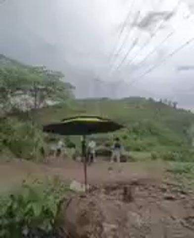 The video begins with a wide shot of a hilly, vegetated area under a cloudy sky. In the foreground, there are people gathered around a large umbrella. The camera then pans to the right, revealing a hillside with power lines stretching across the frame. Smoke begins to rise from the hillside. As the video progresses, the smoke becomes thicker, obscuring part of the hillside and some structures that appear to be buildings. There are multiple plumes of smoke, suggesting a significant fire. The overall environment appears to be rural or semi-rural, with natural vegetation and some evidence of human presence in the form of the umbrella, people, and structures. The sky is overcast, indicating potential for rain or subdued lighting.