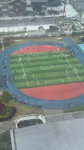 An aerial shot displays a football field encircled by a running track filled with people. The football field is painted with alternating stripes of green and features white lines marking boundaries and goals. People are scattered across the field and along the track. The running track is predominantly red with blue edges. Buildings are visible in the background, along with some roads and minimal traffic. A white roof is partially visible in the foreground.