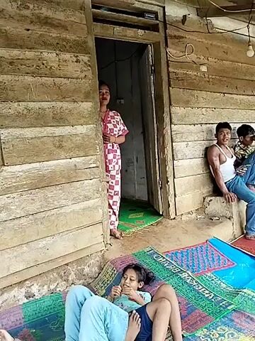 The video shows a group of people gathered outside a wooden house. In the foreground, two young girls are lying on mats on the ground. One girl is wearing a light blue top and pants, and the other is wearing a dark blue top and shorts. Both appear to be looking at something off-camera.

Inside the doorway of the house, a young woman in a pink and white checkered pajama set is peeking out. She is looking towards the camera.

On the steps leading up to the house, a man in a white tank top and blue jeans is sitting, holding a young child. Another man is sitting behind him. Further to the right, another man is visible, seated.

A woman wearing a pink hijab and a floral-patterned dress stands facing the house, with her back to the camera. She appears to be interacting with the people inside or on the steps.

The house appears to be constructed of weathered wooden planks. The ground in front of the house is dirt. Several colorful mats are spread out on the ground. The overall scene suggests a casual, informal gathering.