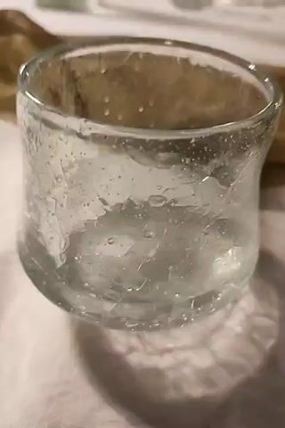 A close-up shot shows a clear glass filled with ice and water. The glass is slightly curved and has a textured surface. The ice appears to be crushed or finely chipped. Bubbles are visible within the water, suggesting it is fizzy or has just been poured. The background is blurred but appears to be a light-colored surface, possibly a tablecloth, with a folded brown fabric item behind the glass. The lighting is soft, casting a subtle shadow of the glass onto the surface. The video pans slightly to the right.