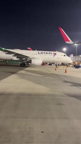 This is a video of a LATAM airplane parked at an airport gate at night. The camera pans across the tarmac, showing the airplane's exterior, including its engines and landing gear. Ground crew members and airport equipment, such as a jet bridge and baggage carts, are visible near the aircraft. The scene is illuminated by airport lights and the airplane's own lights.