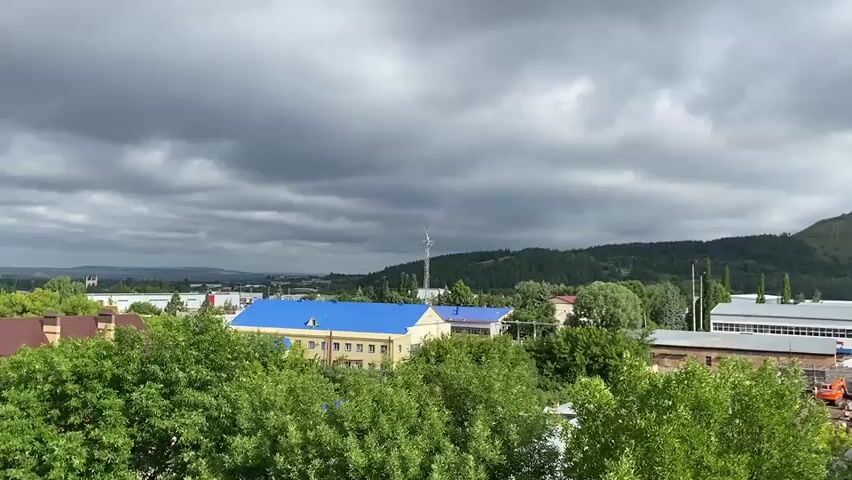 A wide shot shows a town with buildings and trees under a cloudy sky. In the foreground, lush green trees partially obscure the view of a residential area with houses and smaller structures. Behind these, larger industrial or institutional buildings are visible, including one with a bright blue roof and another with a white roof. A prominent hill covered in dense green foliage rises in the background, dotted with several telecommunications towers. The overall atmosphere is overcast but not dark, suggesting a potentially changeable weather day. No people are clearly visible, and no specific actions or interactions are depicted. The scene appears to be a static, daylight view of a semi-rural or industrial town.