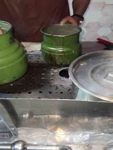 The video captures a close-up of a person, whose hands and torso are visible, preparing a beverage at what appears to be a tea stall or street food vendor in Model Town, Pakistan. The person is wearing a brown long-sleeved garment, possibly a kurta, and a watch on their left wrist.

In the foreground, a stainless steel counter features a perforated section where two large, well-worn, dark green enamel pots are placed. The pot on the left (viewer's left) is larger and appears full. The pot in the center is slightly smaller and sits directly over a small, visible blue gas flame, indicating it is being heated. To the right, a large, lidded silver metallic pot or container rests on the counter.

The primary activity involves the person using a small silver metallic cup to scoop liquid from the larger green pot on the left. They then pour this liquid into the smaller green pot over the flame. Following this, the person uses the same silver cup to stir the contents of the heated pot, suggesting the mixing of ingredients for a hot beverage, likely tea.

The background shows light-colored, possibly hexagonal, wall tiles, hinting at an indoor or semi-enclosed commercial setting. White cloths or rags are visible near the pots, and plastic bags are tucked away under the lower section of the reflective steel counter. A red handle of a utensil can be seen in the upper right background. The general wear and tear on the green pots and the setup are characteristic of traditional tea vendors or chai dhabas common in Pakistan. The time of day is not explicitly clear but artificial lighting suggests it could be evening or night.