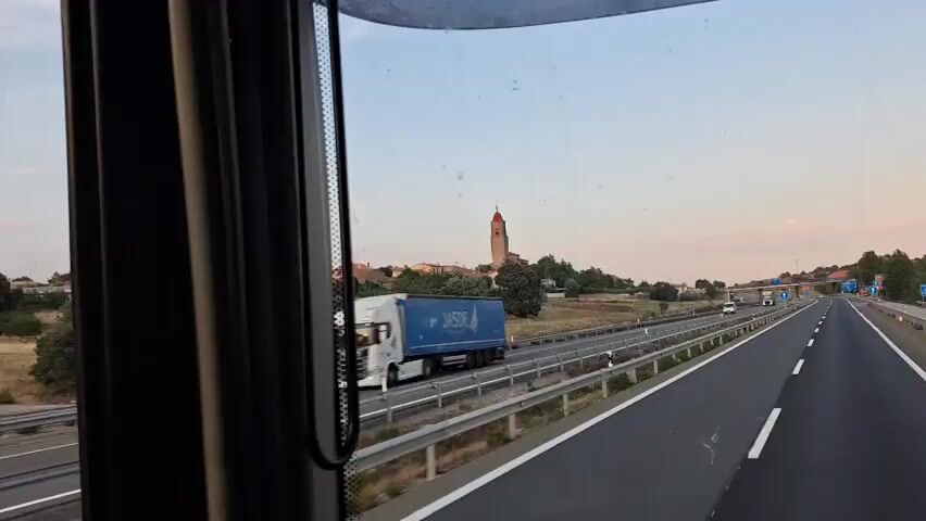 The video captures a view from inside a vehicle, possibly a truck, driving on a highway. The camera is positioned to show the driver's side mirror reflecting the road ahead and the surrounding landscape. A tall tower with a red roof is visible on a hill in the distance, alongside some buildings. The highway has multiple lanes and a metal barrier separates the lanes and the roadside. Several other vehicles, including trucks and vans, are seen on the road. The sky is a soft pastel hue, suggesting it's either sunrise or sunset. The landscape outside the window appears dry and rural, with sparse vegetation and trees. The reflection in the mirror offers a slightly different perspective of the road and the sky.