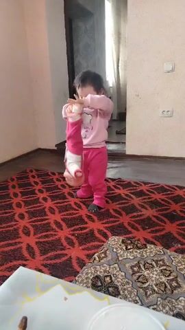 A toddler dressed in a light pink sweater and pink pants stands on a rug and holds a doll aloft. The doll, dressed in similar pink clothing, is positioned upside down. The child appears to be playing with the doll. The room is sparsely furnished, with a patterned rug covering most of the floor, and the walls are painted a neutral color.  A doorway can be seen in the background, along with a table partially visible in the foreground.