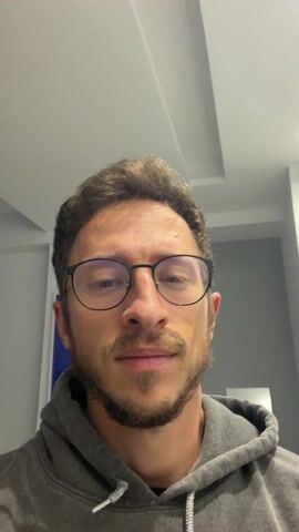 The video begins with a close-up of a man's face. He has brown hair, a short beard, and is wearing glasses and a gray hooded sweatshirt. He says, "Я варю осьминога." which translates to "I'm cooking octopus." The camera then shifts to show a black stovetop with a pot on it. The pot contains an octopus simmering in water with other ingredients like lemon slices and red onion. There are green storage containers on the countertop beside the stovetop.