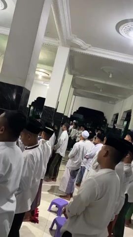 A group of boys, dressed in white shirts and black skullcaps, are standing in a line, facing away from the camera. They are in what appears to be a mosque or a similar religious building. The architecture suggests it's an indoor space with high ceilings and decorative molding. A tall, white column is prominent in the foreground. Other people, also dressed in traditional attire, are visible further back in the hall. The lighting is dim, with some light sources coming from above. The scene appears to be a religious gathering or prayer.
